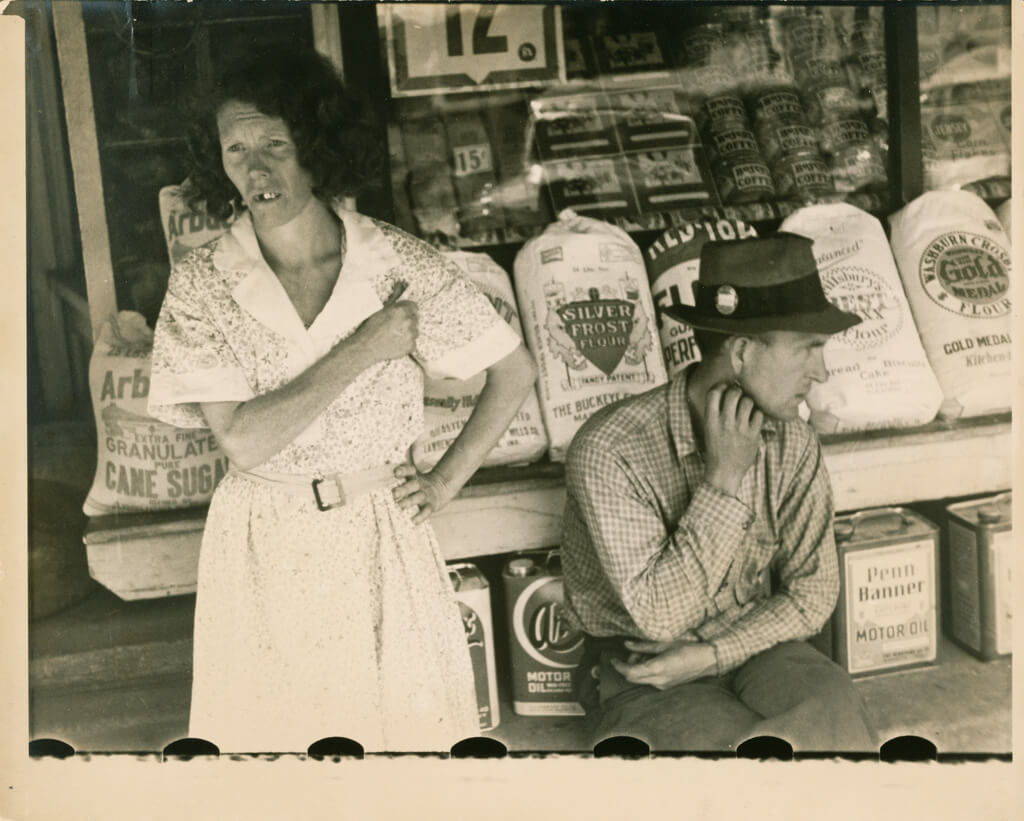 Sepia-toned photograph depicting a middle-aged man and woman sitting in front of a grocery store, looking away from the camera.