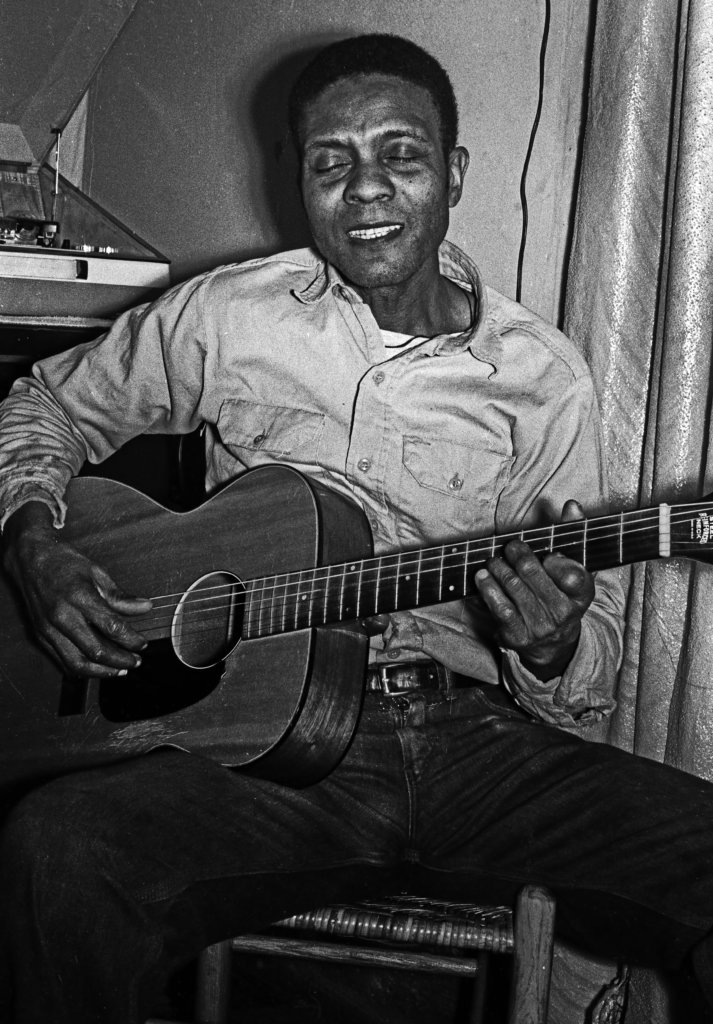 George Henry Bussey holding a guitar and singing with a smile.