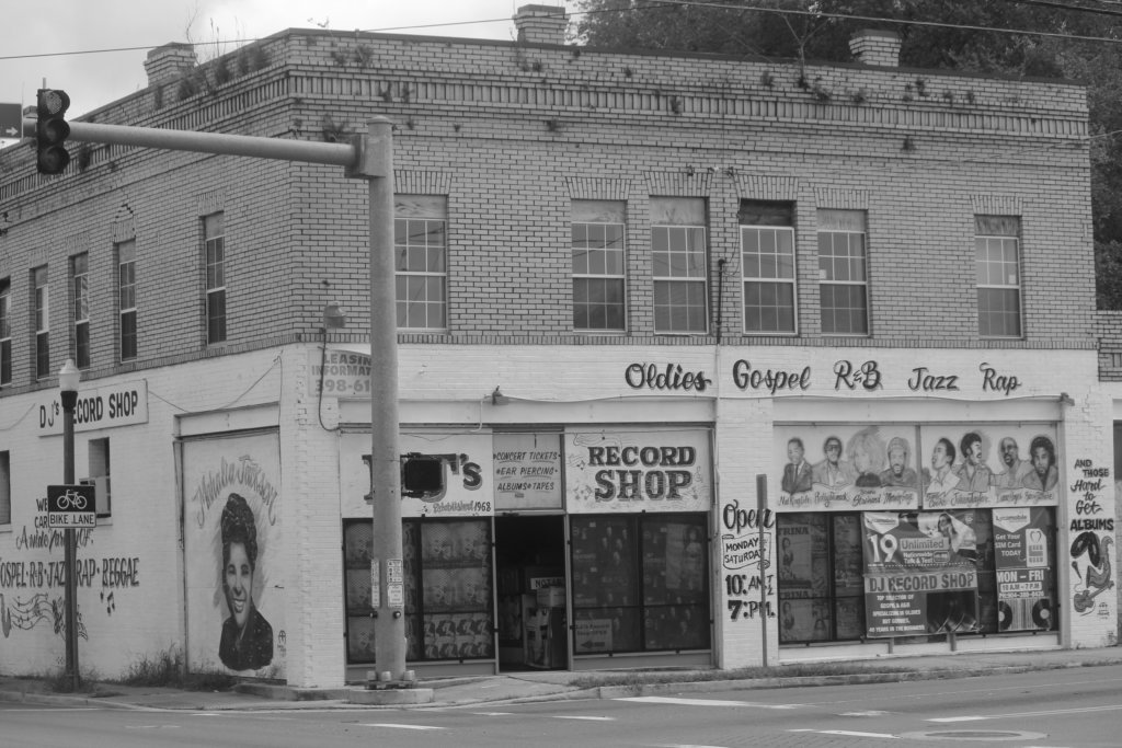 Black and white photograph of storefront of DJ's Record Shop. The words "Oldies," "Gospel," "R&B," and "Rap" are painted in cursive on the building.