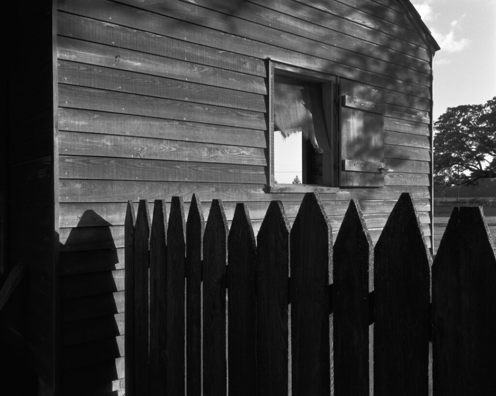 The side of a cabin with an open window stands behind a small strip of fence posts.